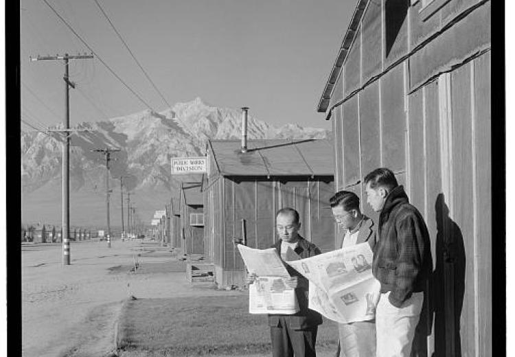 Three men, two reading newspapers, standing in front of Office of Reports, Free Press. Mountains in distance.