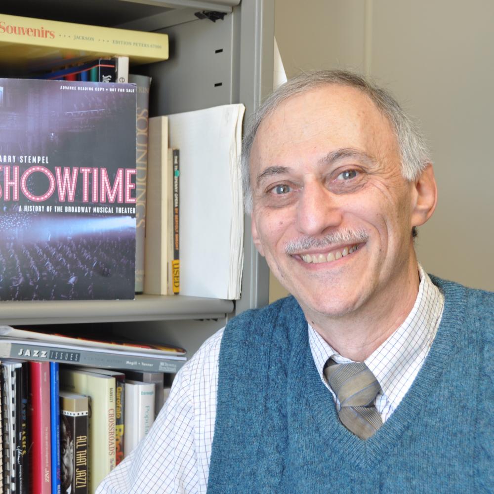 Color photo of Larry Stempel next to a bookcase, wearing a dark green vest, shirt, and tie.