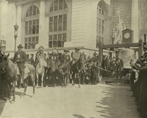 Men on horseback including two with Plains-style feather bonnets in front of a white building