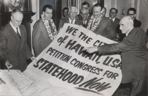 Black and white photograph of 11 men standing around a giant paper that reads, “WE, THE CITIZENS of HAWAII, U.S.A., PETITION CONGRESS FOR STATEHOOD NOW.” Three of the men, including Richard Nixon, are wearing leis.