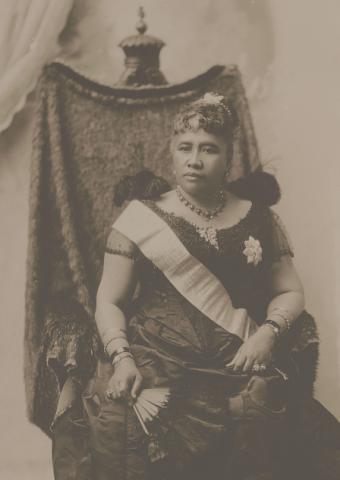 Sepia-toned photograph of Queen Liliʻuokalani seated on a throne, wearing a gown and sash, holding a fan.
