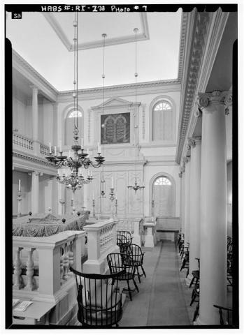 Black and white photo. Interior photo of the Touro Synagogue. Showcases large white columns, and black chairs surrounding the bimah, an elevated space used to read the Torah. 