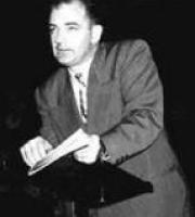 McCarthy in a dark suit, leans on a small podium