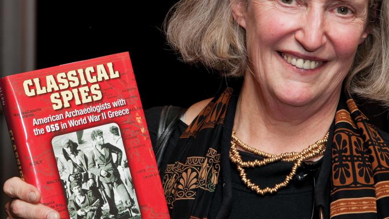 Heuck Allen in a striped scarf, posing with her book "Classical Spies"