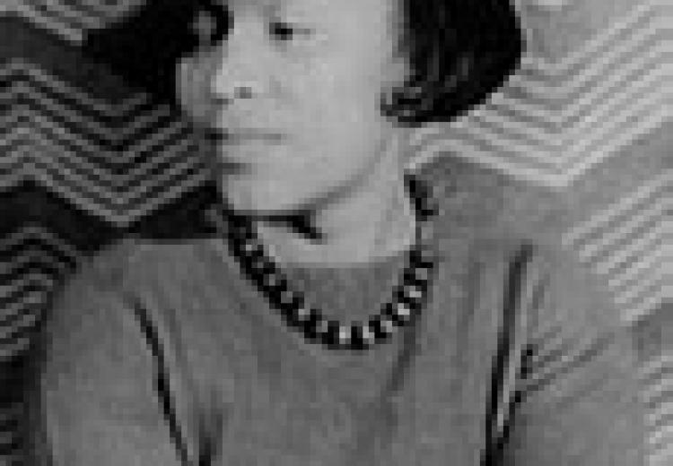 Zora Neale Hurston, in a black hat, grey sweater, and beaded necklace, face turned slightly to her left