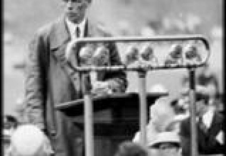 lindbergh, in a long overcoat and suit, stands at a podium on a stage, in front of a crowd of people