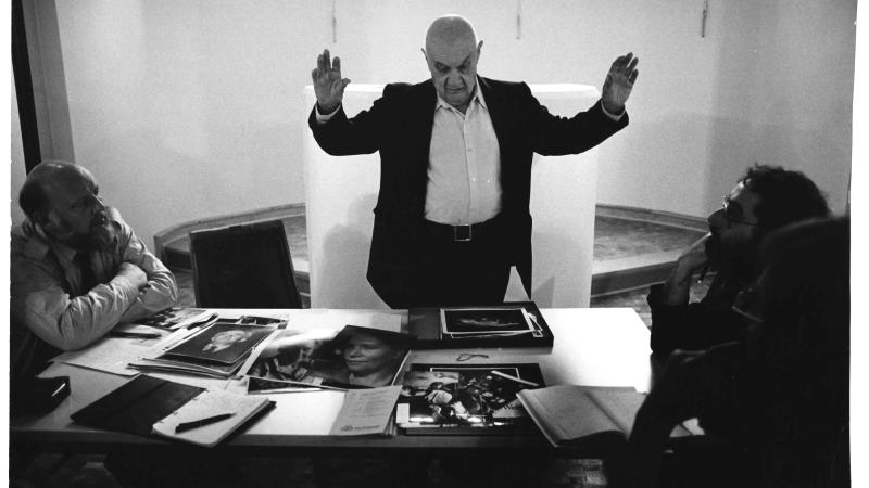 Roman Vishniac stands with arms outstretched looking at photographs on a table