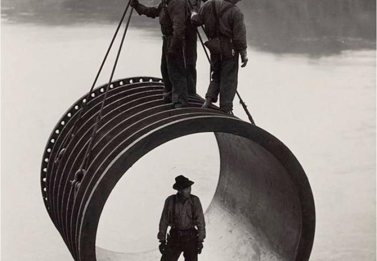 Four men, three standing on top and one standing inside, riding on large casing section of pipe suspended by cable as it is moved into position by a crane, reservoir in the background during construction of the Grand Coulee Dam, Washington