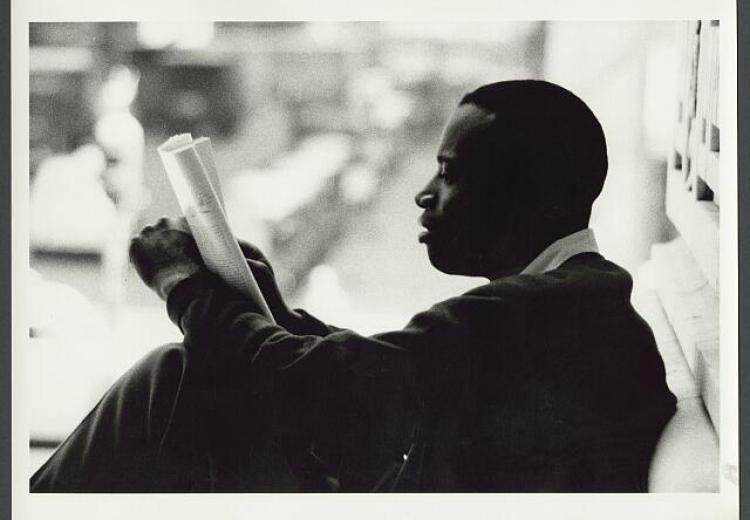 Photograph shows a man, from the side, sitting against a wall and reading a newspaper.